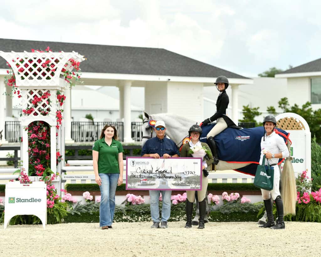 Five people and a horse pose outdoors with a large check and ribbon after an equestrian competition. A woman on horseback is draped in a blue blanket. Flowers and sponsor banners are visible.