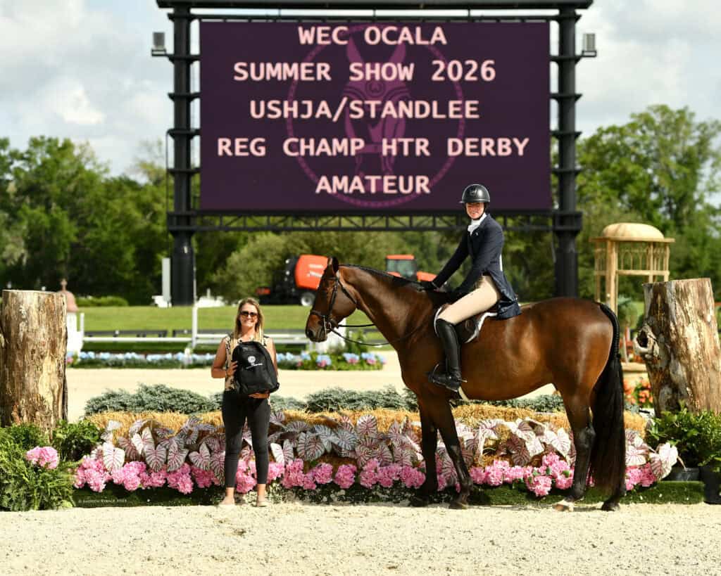 A rider on horseback and a woman standing beside the horse pose in front of a sign reading "WEC Ocala Summer Show 2026 USHJA/Standlee Reg Champ HTR Derby Amateur," with flowers in the foreground.