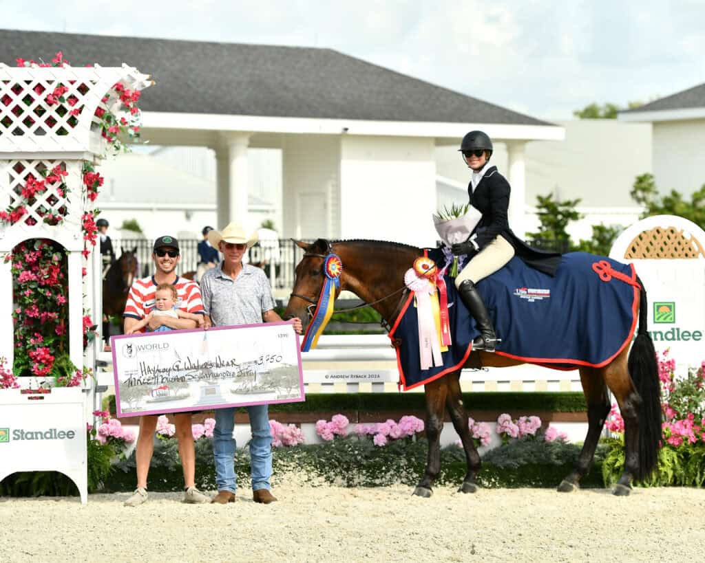 A rider on a horse wearing a champion's blanket and ribbons poses with two men holding a large prize check at an outdoor equestrian event.