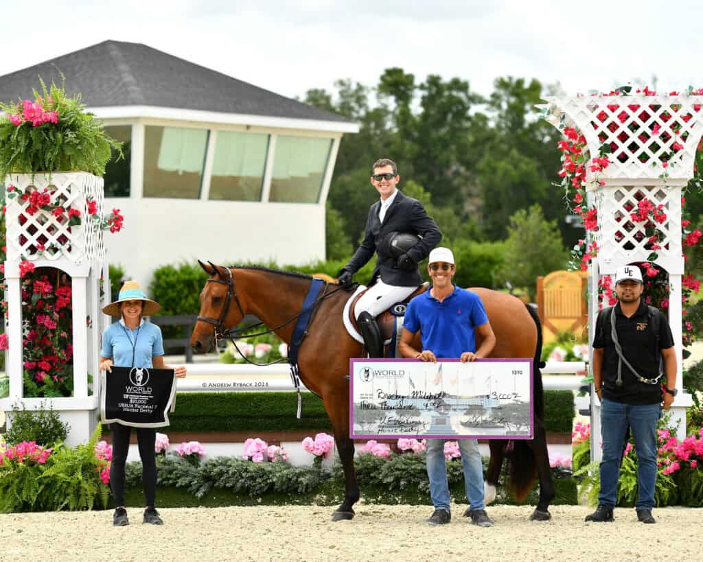Four people and a horse pose at an award ceremony; one person on horseback, two holding a large check, and one holding a tote bag, with flowers and white lattice structures in the background.