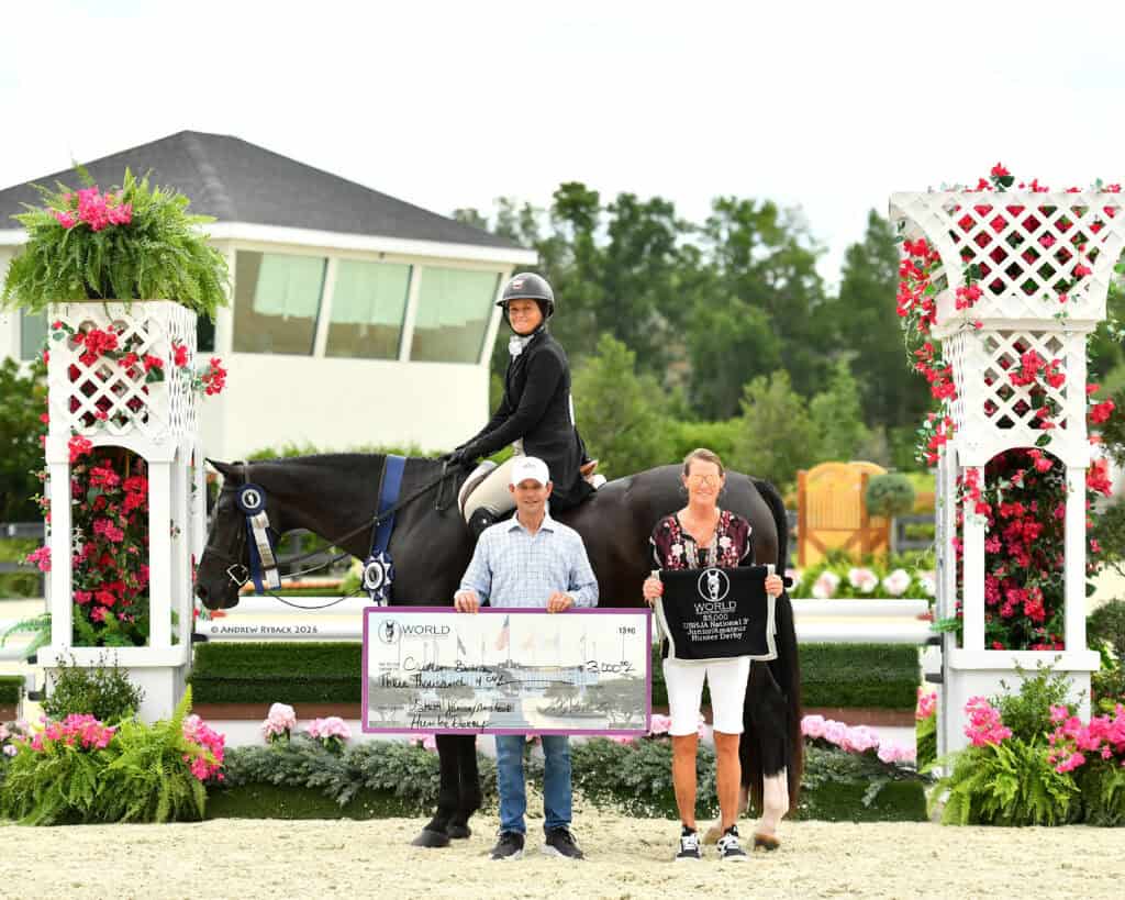 Three people pose with a horse at an outdoor equestrian event, holding a large check and a banner, with flower arrangements and greenery in the background.