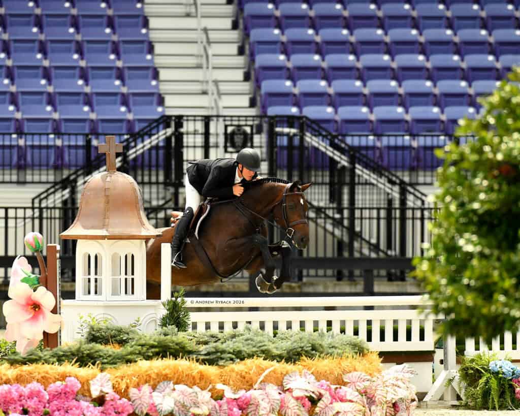 A rider in formal attire guides a horse over a jump decorated with flowers in an empty arena with rows of purple seats.