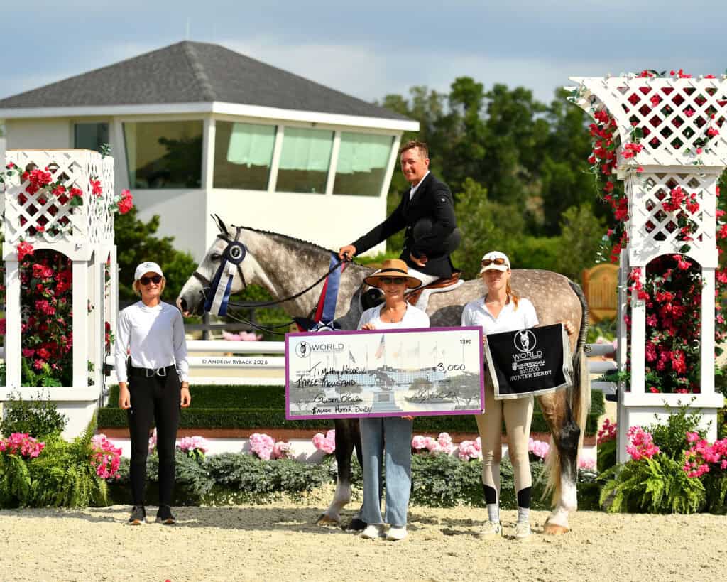 A man on a horse poses with three women, one holding a large check, in an outdoor equestrian arena with flowers, white fencing, and a modern building in the background.