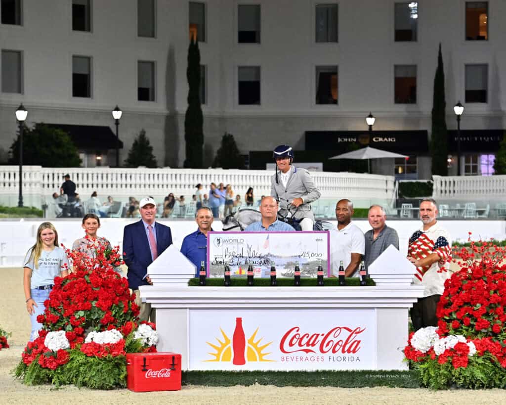 A group of people stands around a large check and Coca-Cola display at an outdoor event, with a man in equestrian attire sitting on a horse behind the table.