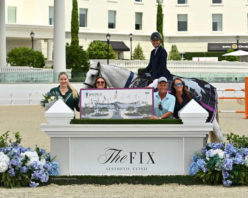 A rider on a gray horse holds a large ceremonial check, posing with four people behind a white display labeled "The Fix Aesthetic Clinic" at an equestrian event.