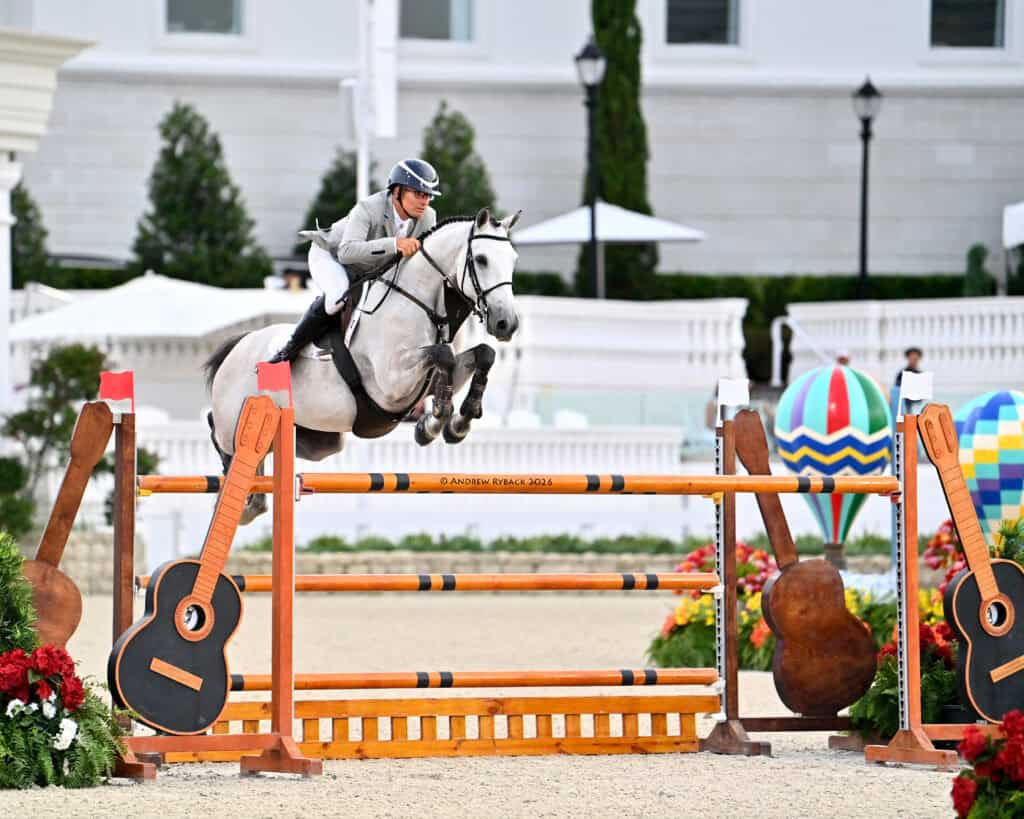 A rider in a helmet and jacket guides a gray horse over a high show jumping obstacle decorated with large guitar shapes in an outdoor arena.