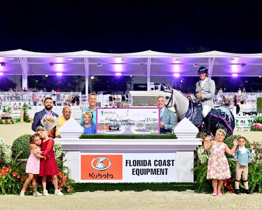 A group of people, including children, stand in front of a show jumping prize sign with a man on a gray horse holding a large check at an outdoor event.
