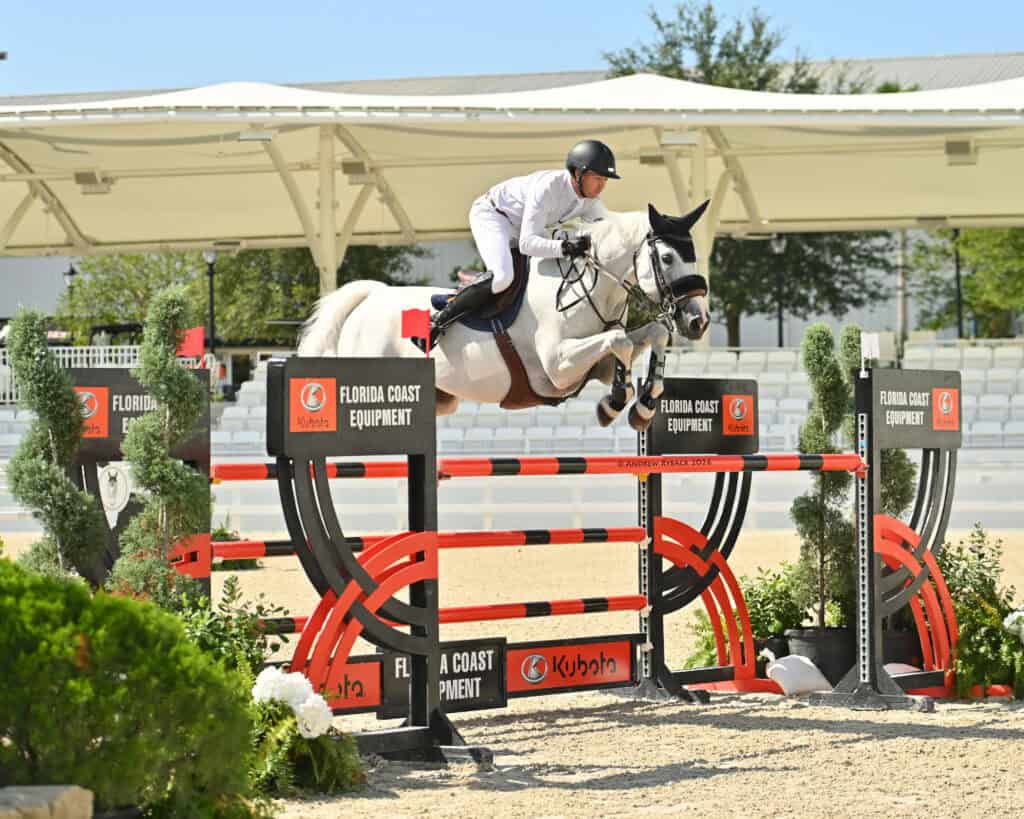 A rider in white jumps a gray horse over a red and black obstacle at an outdoor equestrian event.