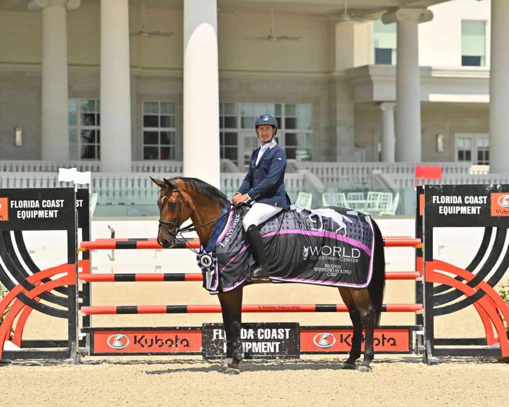 A rider in formal attire sits on a horse draped in a winner’s blanket in front of a show jumping obstacle at an equestrian event.