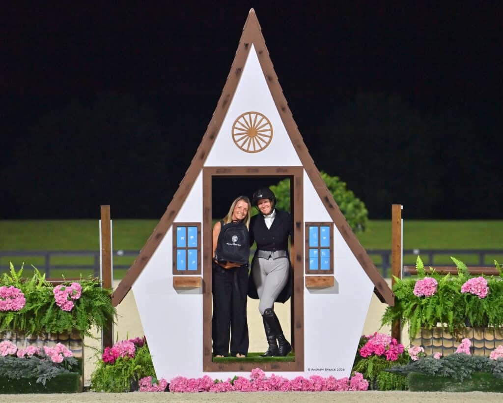 Two women stand smiling inside a decorative house-shaped frame, one in equestrian attire and the other holding a prize bag, surrounded by flowers and greenery at night.