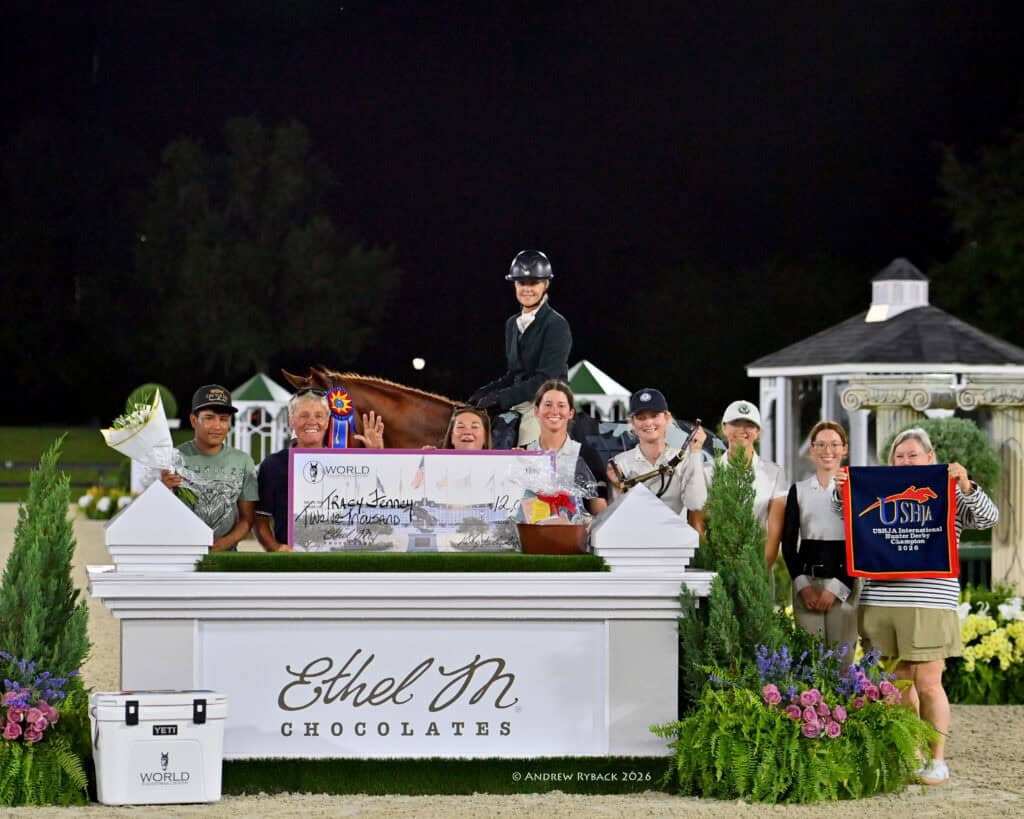 A group of people pose around a horseback rider holding a ribbon, behind an Ethel M. Chocolates sign at an equestrian event.