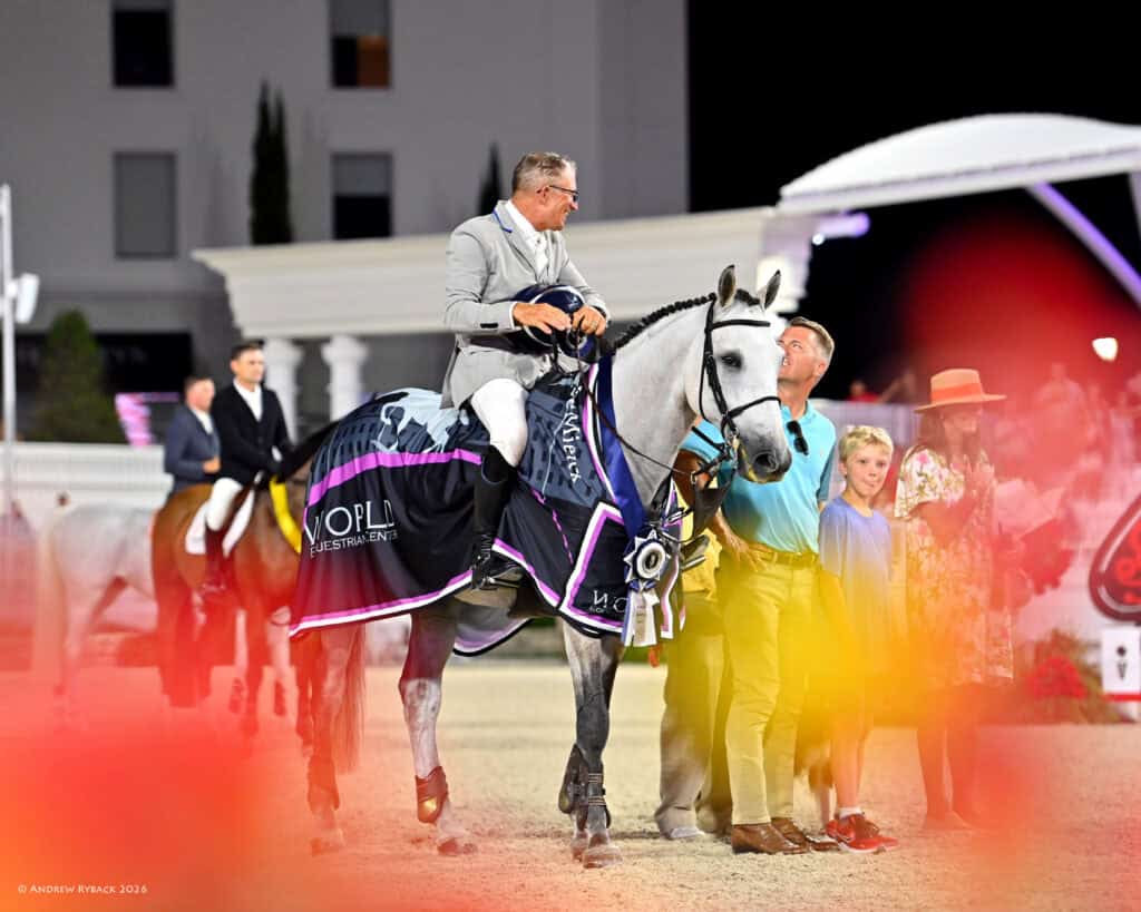 A man in a light suit sits on a decorated horse with a winner’s ribbon, surrounded by people, during an equestrian event at night.