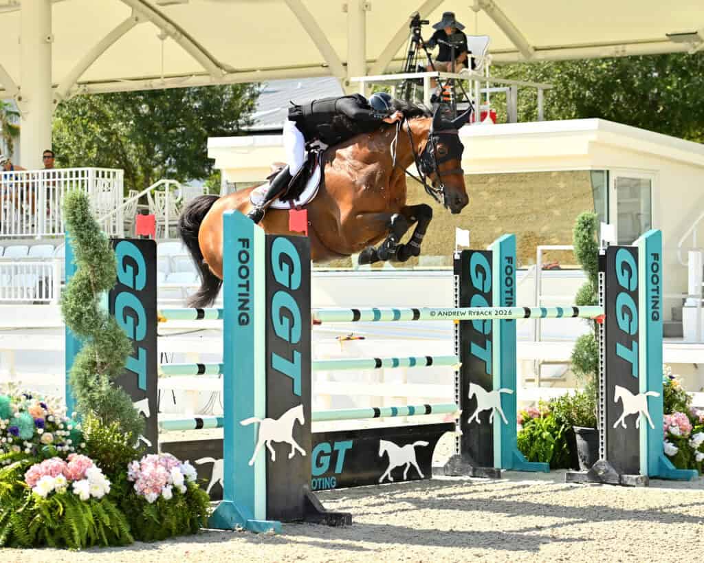 A rider and horse in mid-air jump over a hurdle during an equestrian competition, with GGT branding and a judge seated in the background.