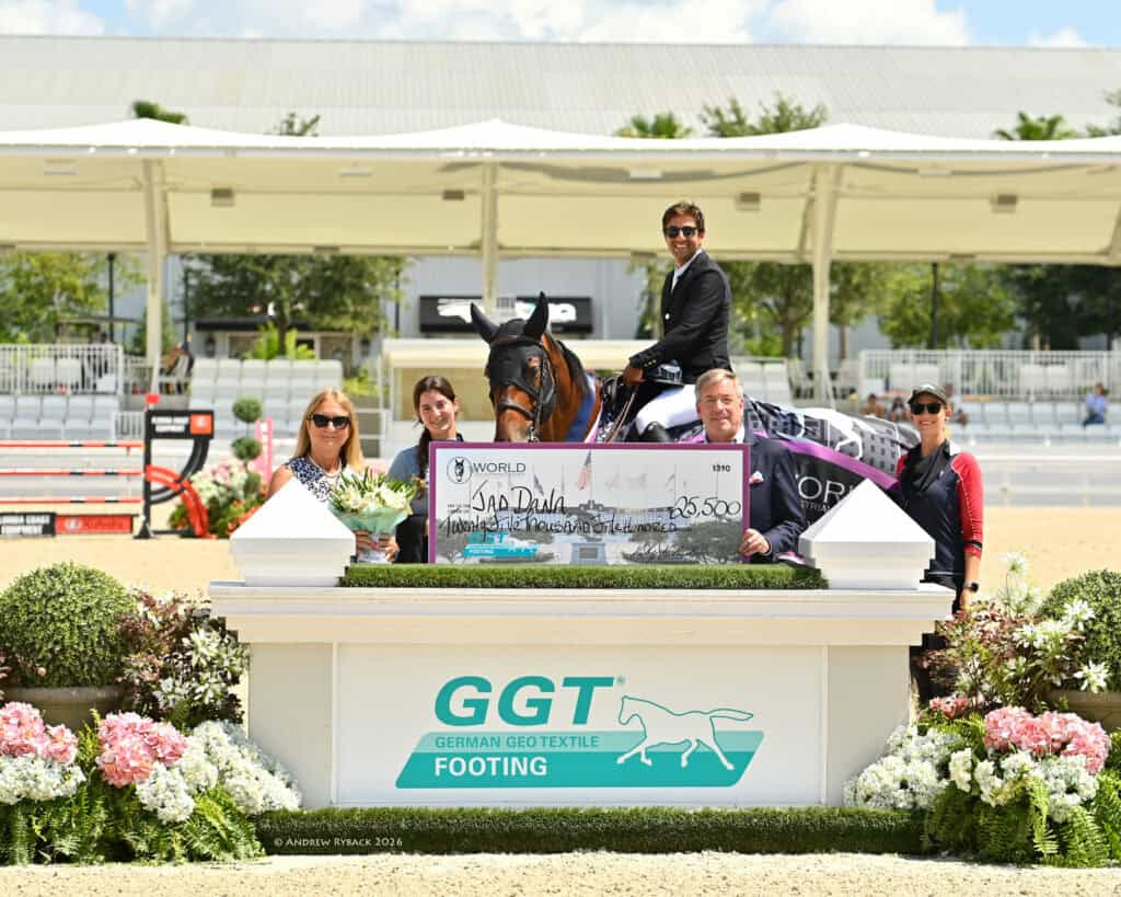 A group of five people and a horse pose outdoors with a large check for $25,500 at an equestrian event. Flowers and event signage are displayed in front of them.