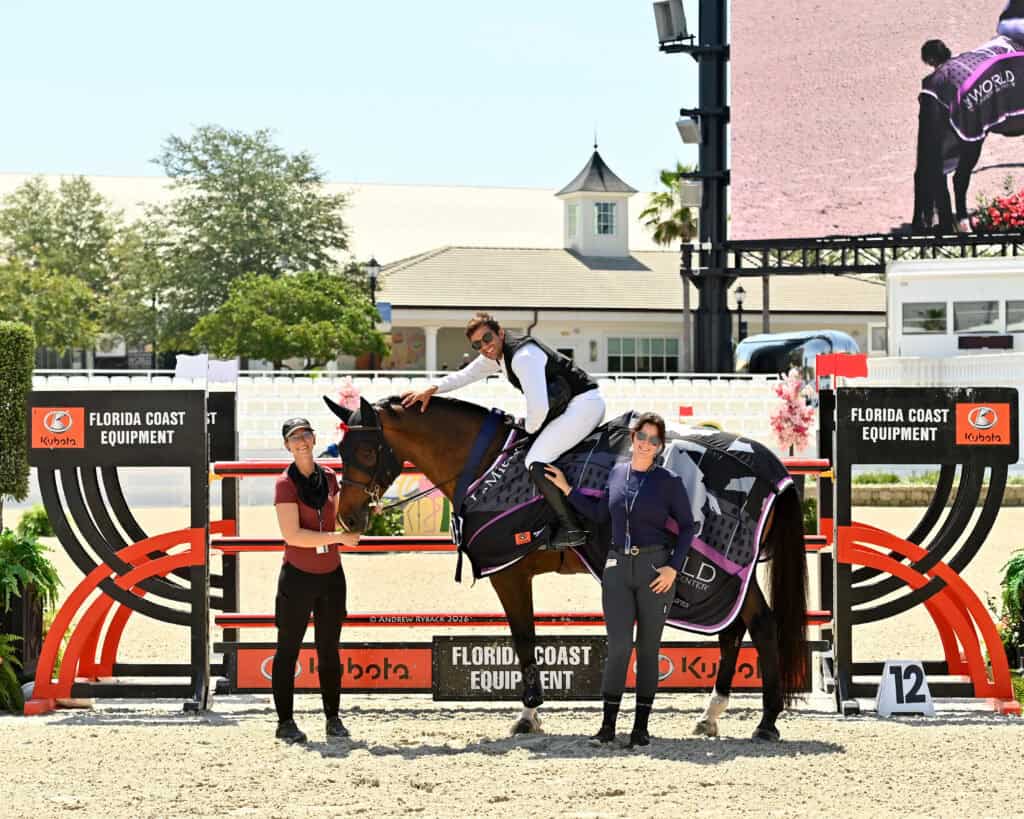 Three people pose with a horse in front of a Florida Coast Equipment jump at an outdoor equestrian event; one person is mounted, the other two stand on either side.