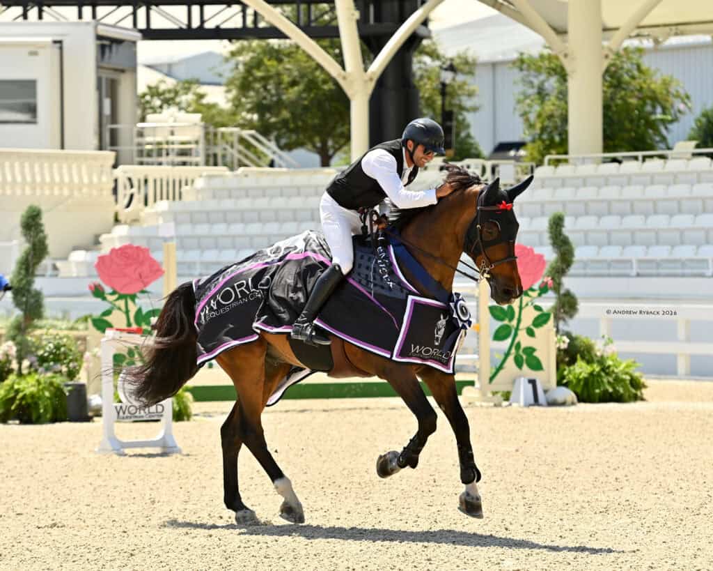 A rider in equestrian attire leans forward on a horse draped in a winner’s blanket, celebrating in an outdoor arena with empty stands and decorative elements.