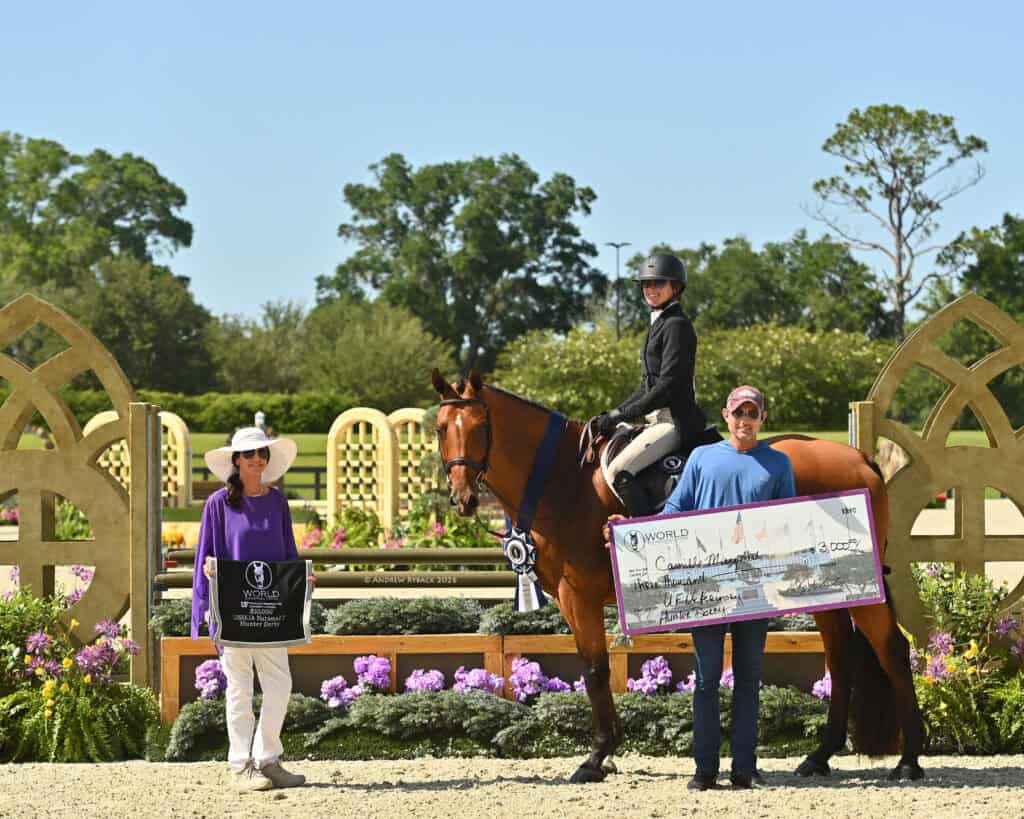 Three people and a horse at an equestrian event; one person is mounted, while the others hold a plaque and a large check in front of a decorated jump and flower arrangements.