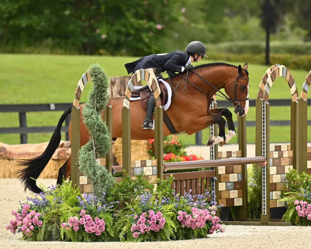 A rider in formal attire and helmet guides a bay horse over a jump during an equestrian event, with flowers and greenery in the foreground.