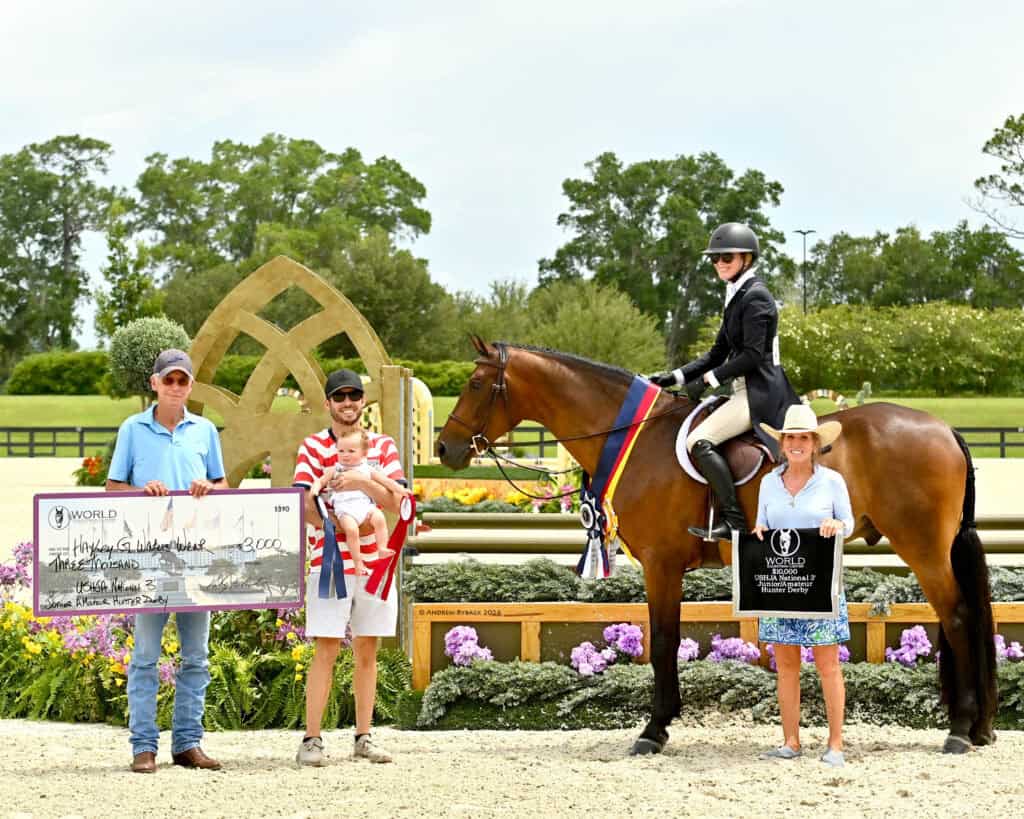 Four people and a horse pose at an outdoor equestrian event. One person holds a large check, another holds a baby, and the rider displays awards. Flowers and banners are visible.