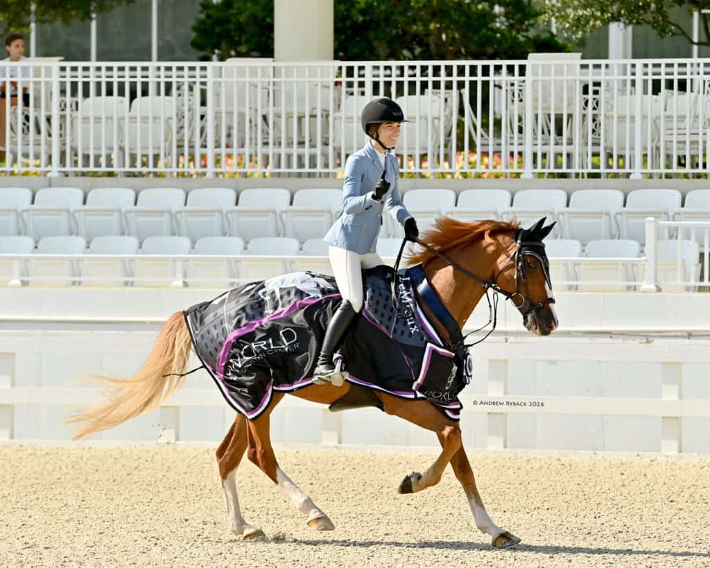 A rider in a light blue jacket and helmet rides a chestnut horse draped in a winner’s blanket in an empty arena, holding up a finger in a "number one" gesture.