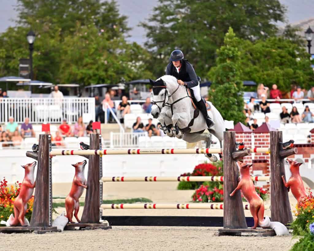 A rider on a gray horse jumps over a tall obstacle decorated with fox statues during an equestrian event, with spectators watching in the background.