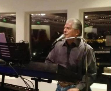 An older man plays a keyboard and sings into a microphone in an indoor venue with large windows and string lights.