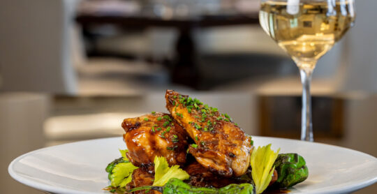Glazed chicken pieces with chives and green peppers on a white plate, with a glass of white wine in the background.