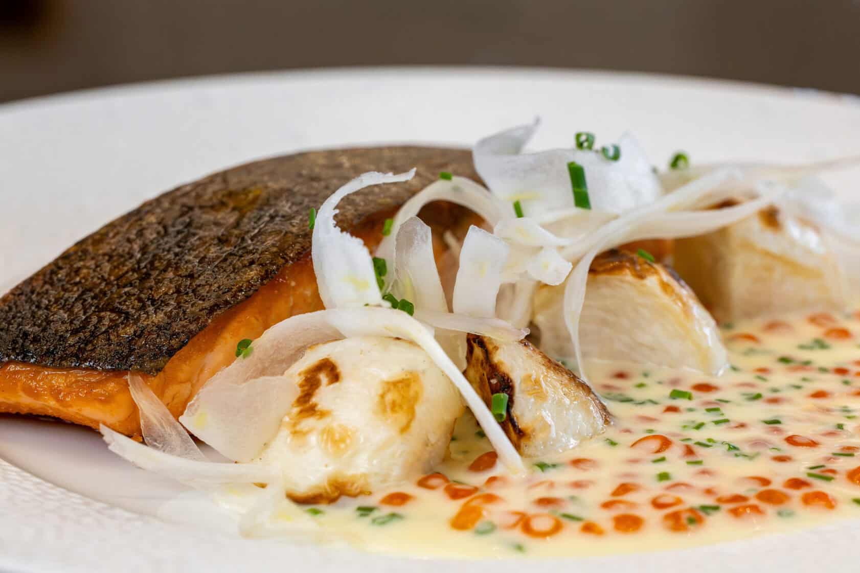 A plated dish with seared salmon fillet, roasted vegetables, thinly sliced onions, and creamy sauce with herb and orange oil garnish.