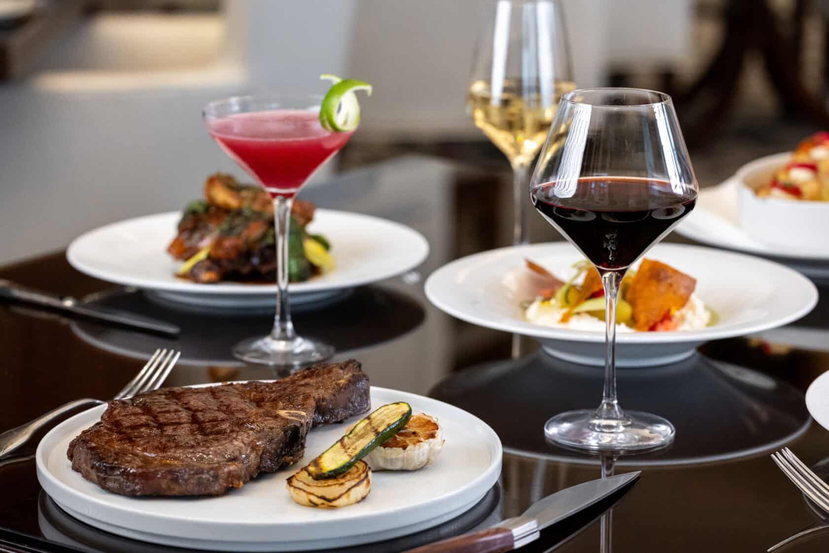 A steak with grilled vegetables and a glass of red wine on a table, accompanied by a pink cocktail and plates of various dishes in the background.