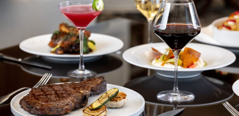 A plated steak with grilled vegetables, a red wine glass, and a pink cocktail on a table with other plated dishes in the background.