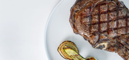 A grilled steak with seared grill marks is served on a white plate alongside roasted slices of zucchini and onion.
