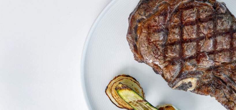 A grilled steak with seared grill marks is served on a white plate alongside roasted slices of zucchini and onion.