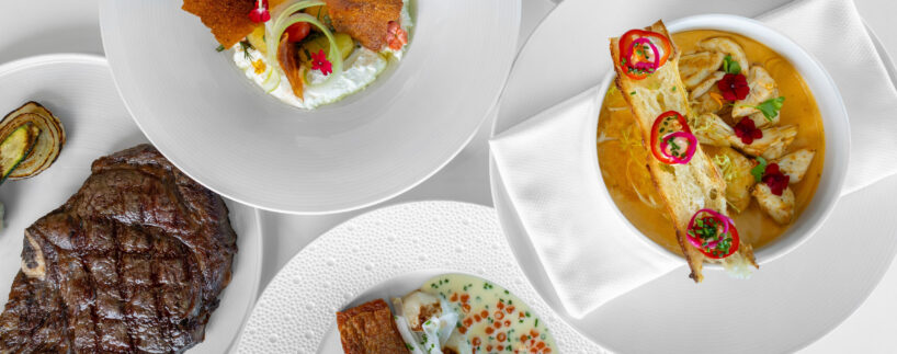 Five white plates with various gourmet dishes, including grilled steak, soup with pastry, fish in sauce, lamb shank with greens, and a salad with crispy elements, arranged on a white table.