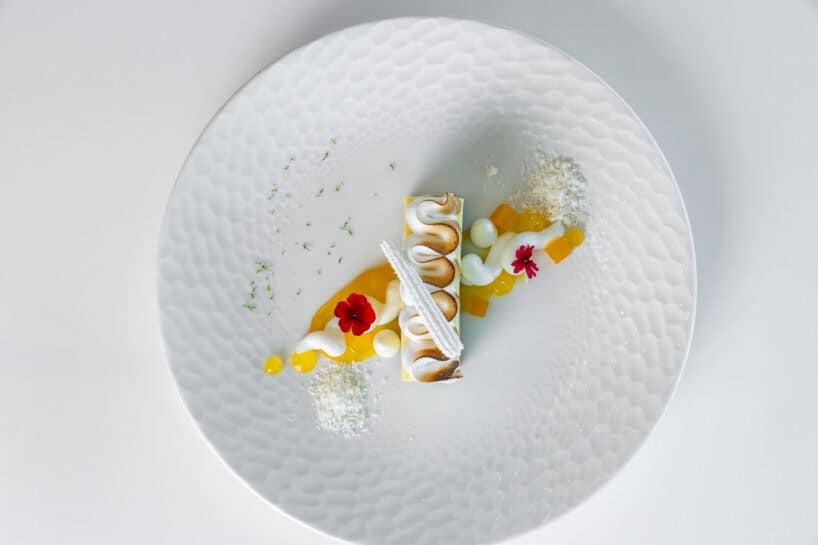 A plated dessert features a rectangular pastry with piped cream, yellow sauce, white crumbles, red edible flowers, and green garnish on a textured white plate.