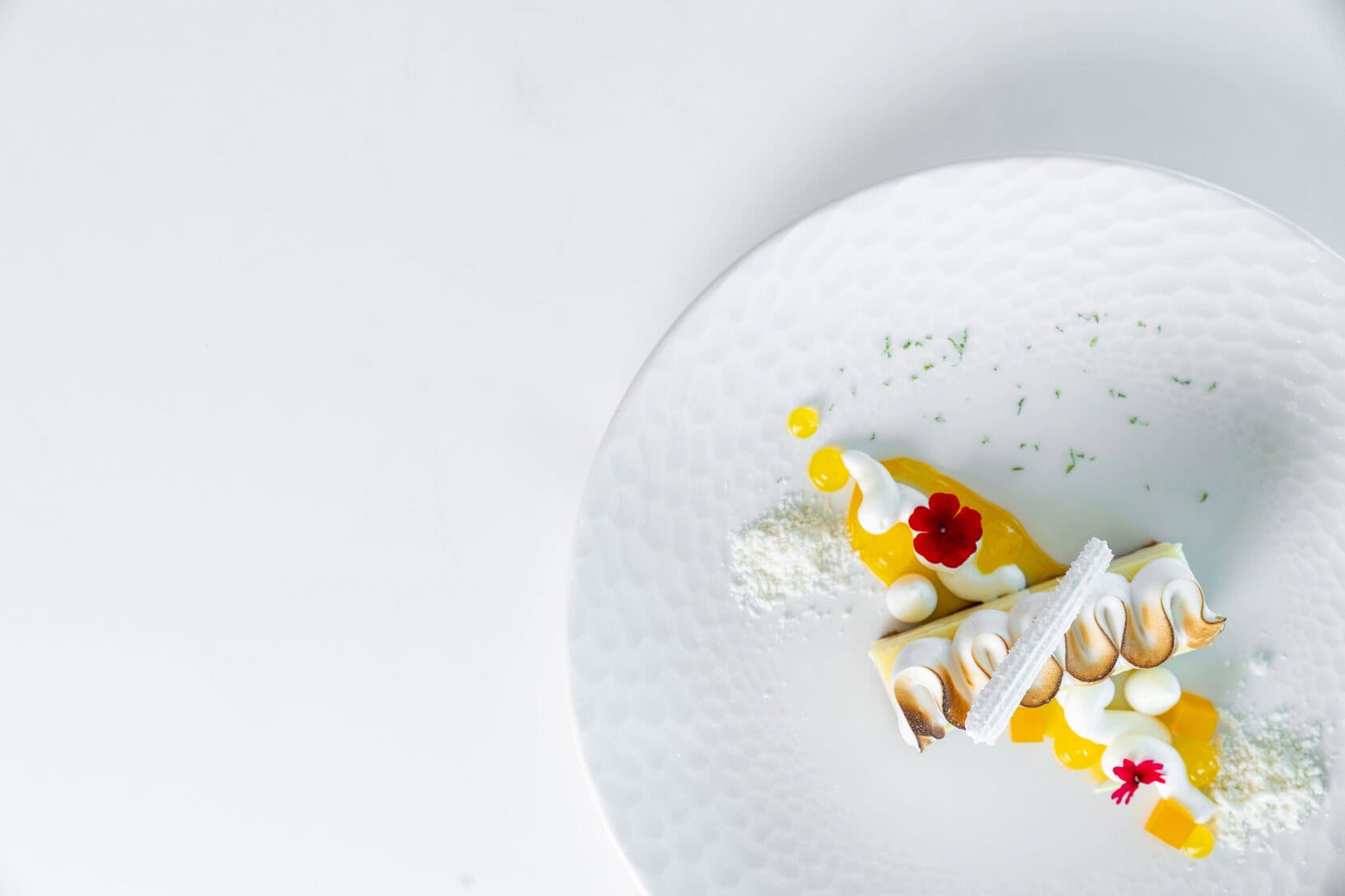 A slice of lemon meringue pie with decorative cream, a red edible flower, and citrus sauce on a white textured plate.