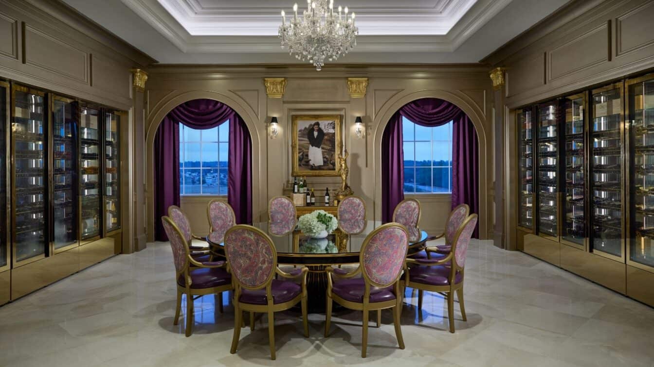Elegant dining room with a round table, eight ornate chairs, chandelier, floor-to-ceiling wine storage, purple curtains, and a large portrait on the back wall.