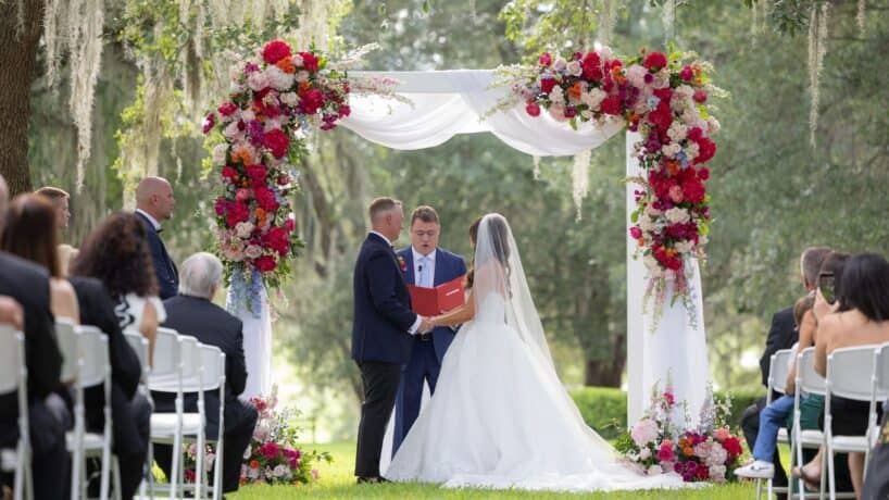 A bride and groom stand under a floral arch exchanging vows during an outdoor wedding ceremony, witnessed by seated guests.