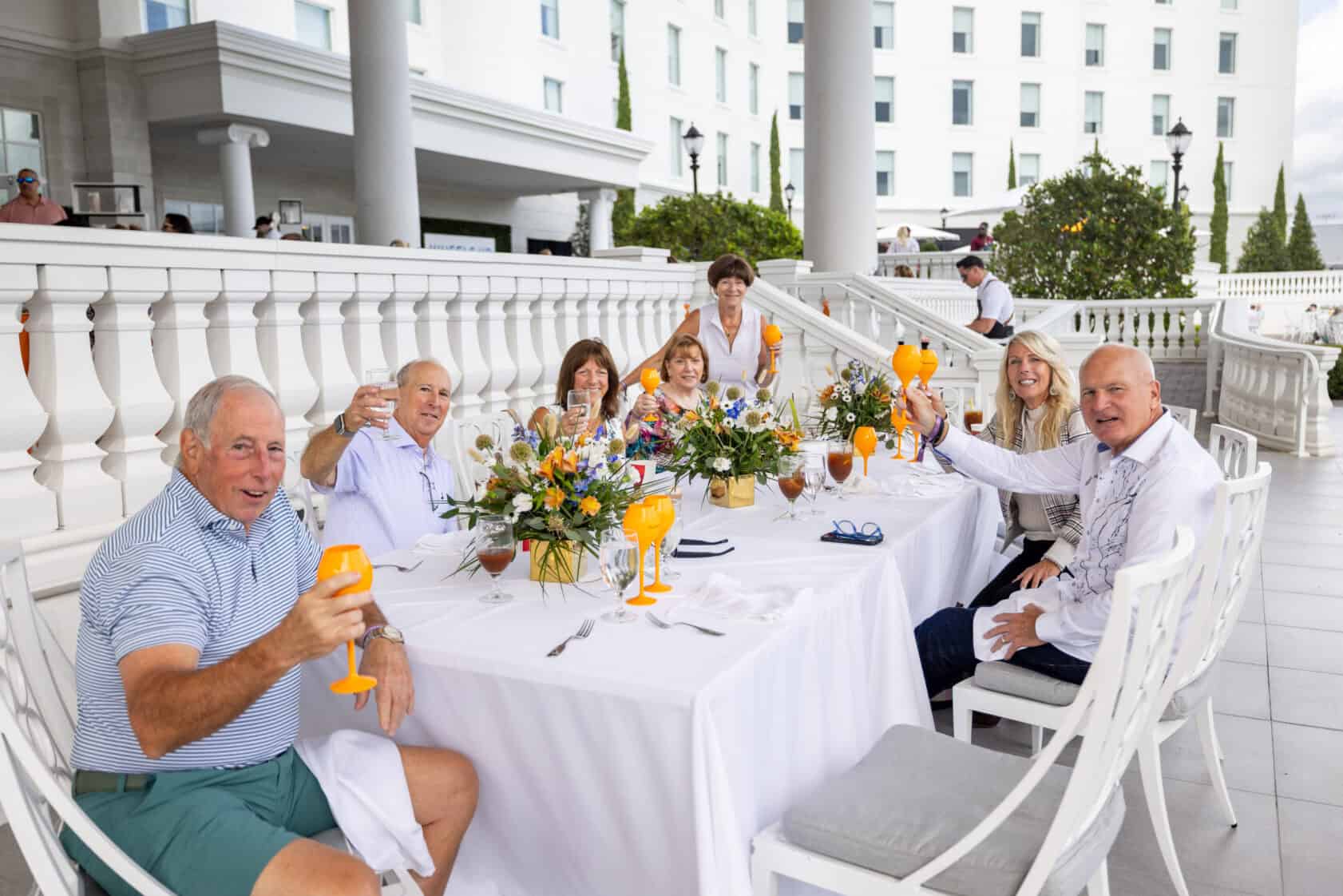 Seven adults sit around a white-clothed outdoor table with drinks raised in a toast, in front of a large white building with columns.