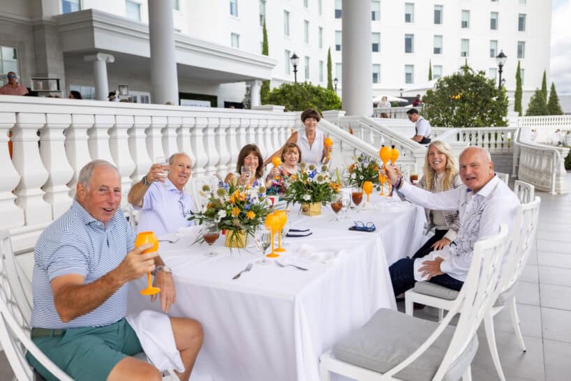 Seven adults sit around a white-clothed outdoor table with drinks raised in a toast, in front of a large white building with columns.