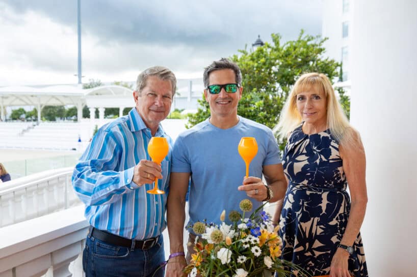 Three adults stand outdoors holding orange drinks, with one man wearing sunglasses. They are next to a flower arrangement and a white building under a cloudy sky.