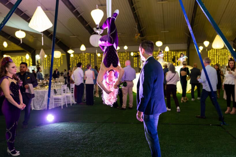 An aerial performer hangs upside down on a hoop, holding a bottle toward a man in a blazer, at an indoor event with guests, festive lights, and decorations.