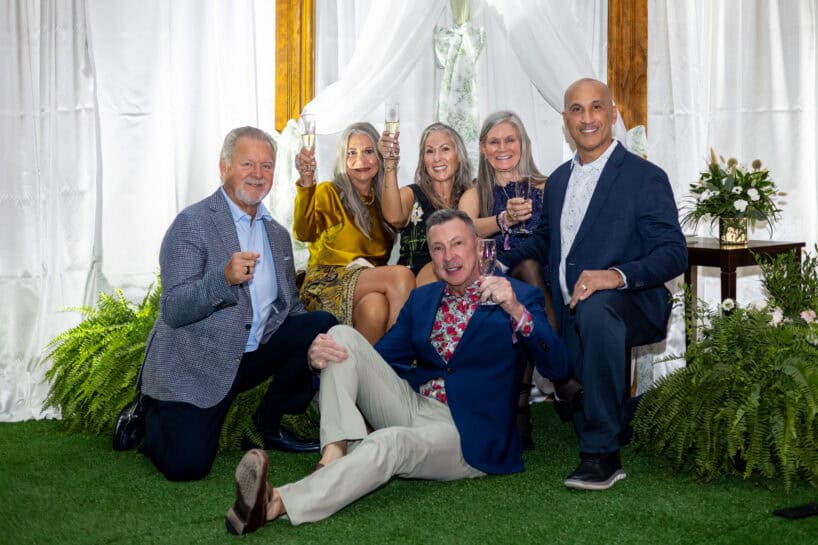 Six adults dressed in semi-formal attire pose together indoors on green turf, holding up glasses in a toast, with white drapery and plants in the background.