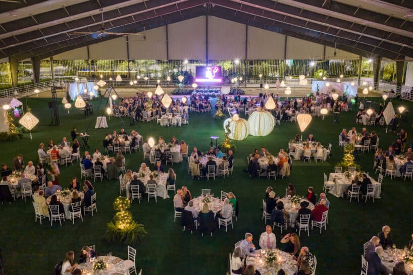 Large indoor event with many round tables, guests dining, and decorative lighting, taking place on a green artificial lawn under a high ceiling.
