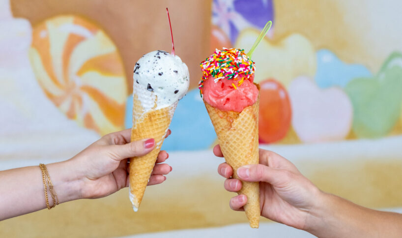 Two hands holding ice cream cones, one with cookies and cream topped with a cherry, the other with pink ice cream and sprinkles, above a pink box of cookies and macarons.