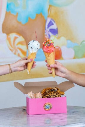 Two hands hold ice cream cones with sprinkles and cherries above a pink box of assorted cookies and macarons on a marble table.