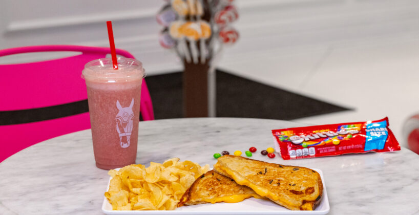A plate with a grilled cheese sandwich and potato chips sits on a white table, alongside a pink smoothie, a pack of Sweetarts, and scattered colorful candy.