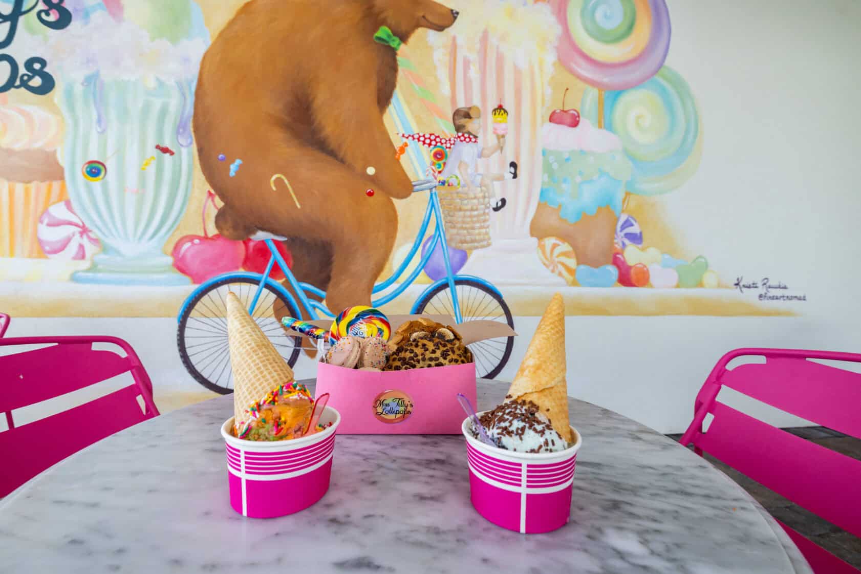 Two pink cups of ice cream with waffle cones and toppings sit on a marble table; a mural of a bear on a blue bicycle decorates the wall in the background.