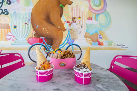 Two pink cups of ice cream with waffle cones and toppings sit on a marble table; a mural of a bear on a blue bicycle decorates the wall in the background.