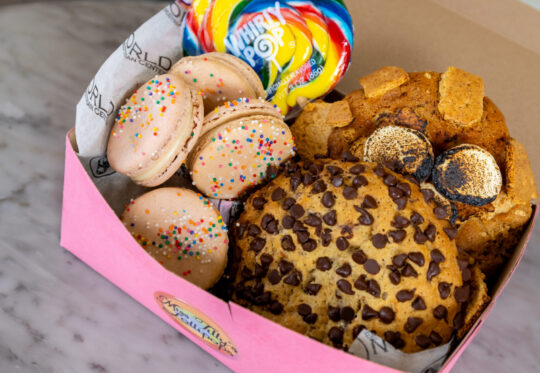 A pink box on a marble surface contains macarons with sprinkles, a large chocolate chip cookie, a rainbow lollipop, and assorted cookie pieces.