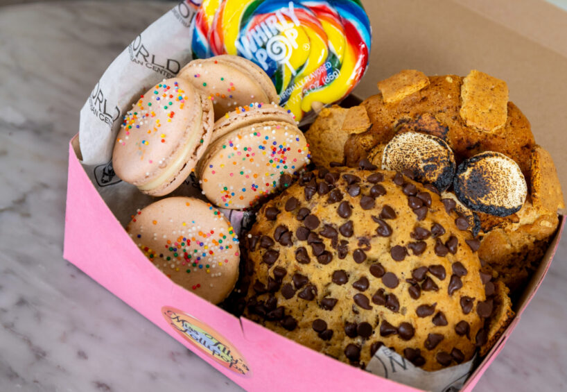 A pink box on a marble surface contains macarons with sprinkles, a large chocolate chip cookie, a rainbow lollipop, and assorted cookie pieces.